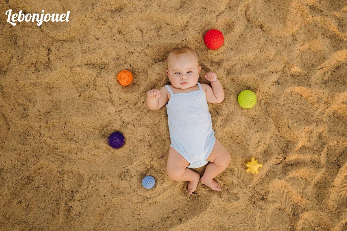 Éveil sensoriel en douceur : bébé explore les textures avec des balles colorées sur le sable.
