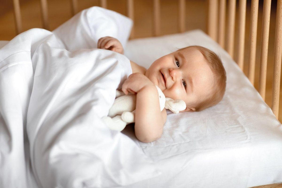 Un bébé souriant tenant son doudou dans un berceau en bois, symbole de confort et de lien affectif.