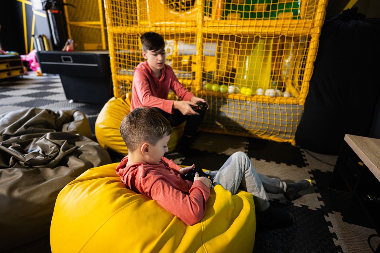Deux frères jouent à la console dans un espace enfants, sur un pouf jaune confortable.