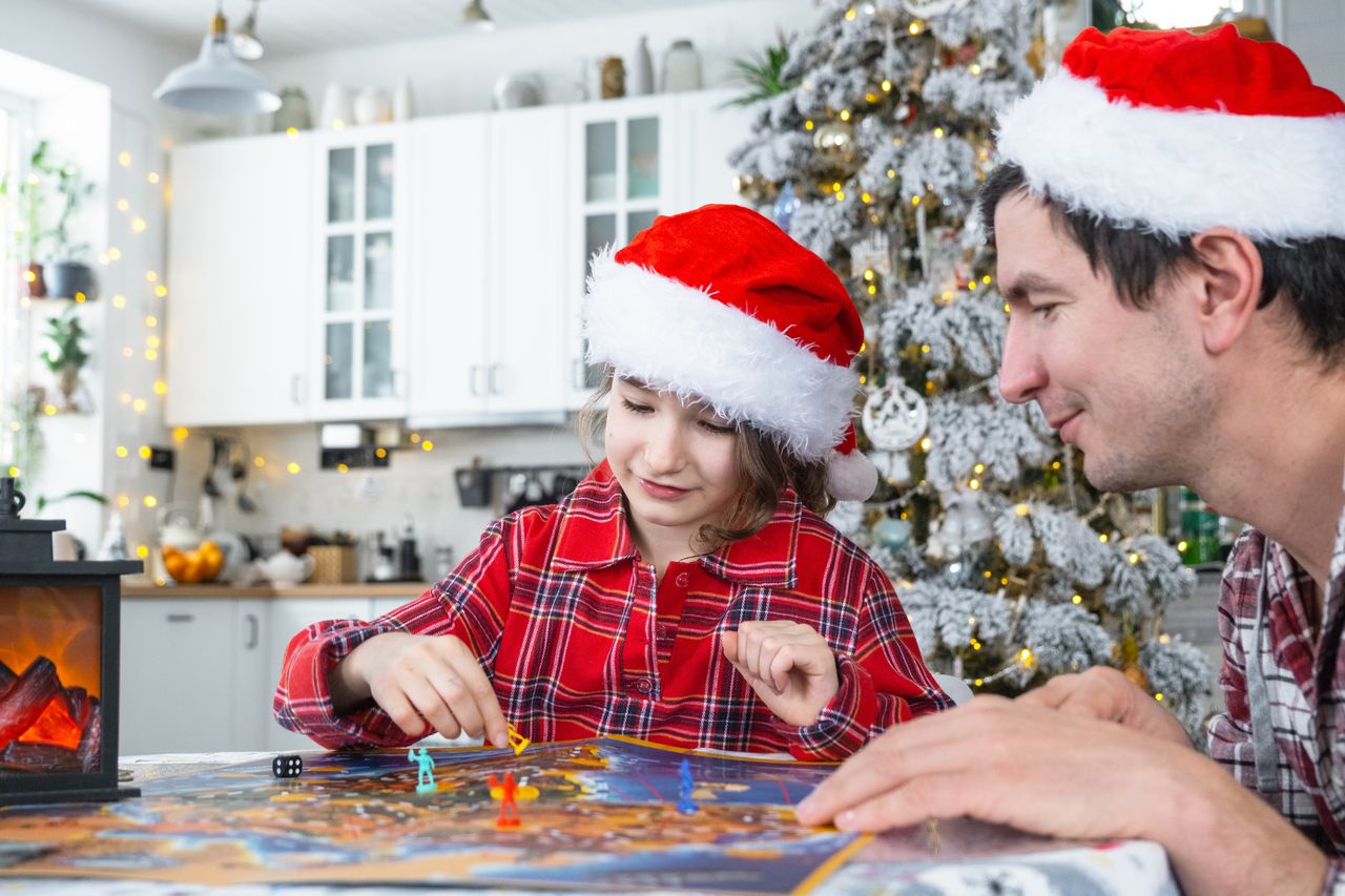 Un père et sa fille en bonnets de Père Noël jouent à un jeu de plateau devant le sapin.