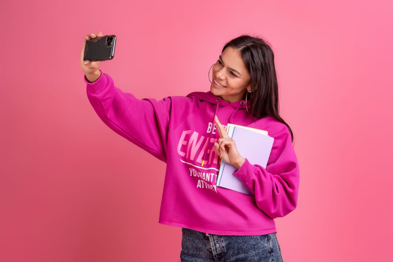 Jeune femme souriante en hoodie rose, avec carnets et smartphone, dans un style tendance et féminin.