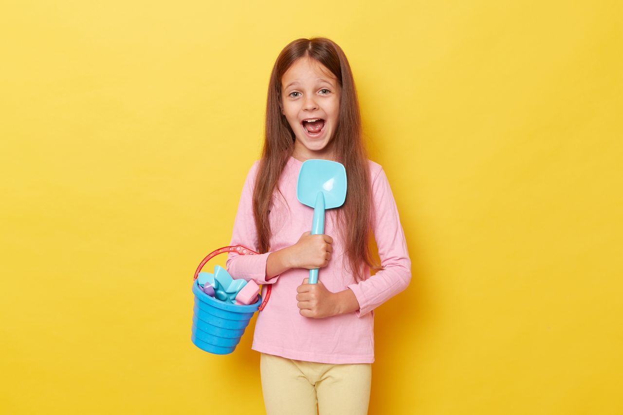 Jeune fille joyeuse avec seau et pelle de plage, jouets colorés sur fond jaune estival.