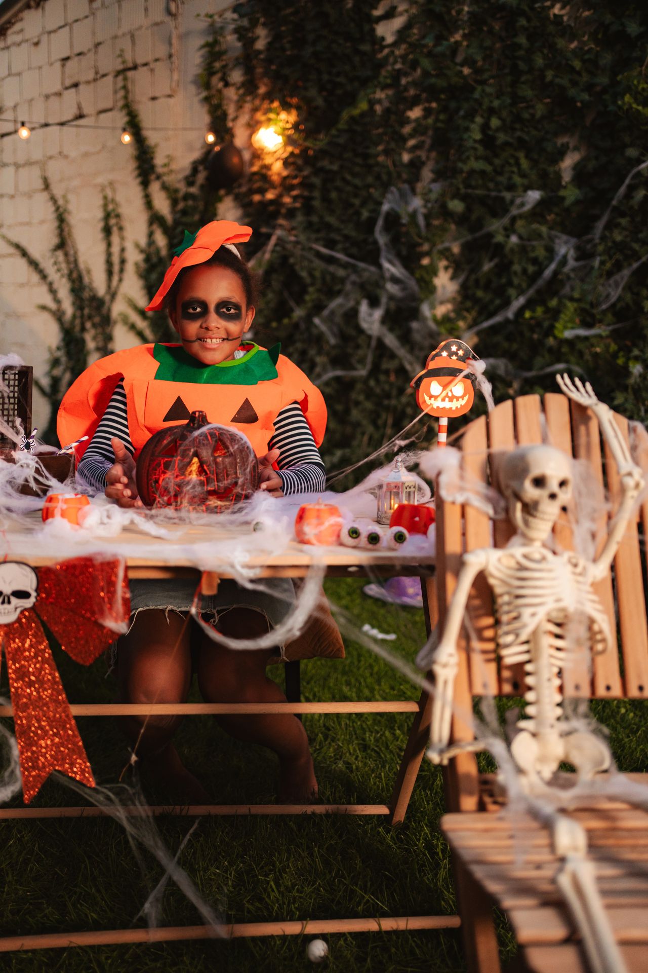 Petite fille maquillée en citrouille pour Halloween, tenant une lanterne-jack-o'-lantern dans une ambiance familiale.