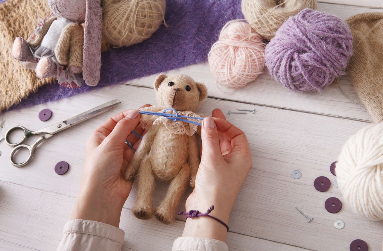 Mains d’une couturière qui assemble un ours en peluche, entourée de pelotes de laine.